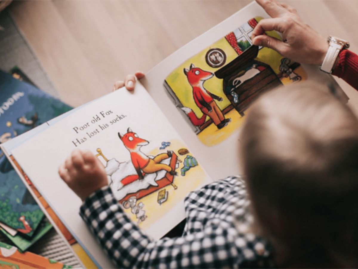 Toddler holding an open picture book with an adult hand flipping the page.