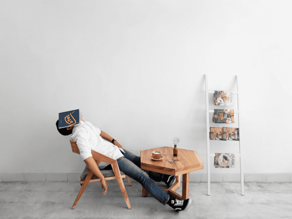 Man sitting on a wooden chair with a coffee. He has an open book over his face and appears to be sleeping.