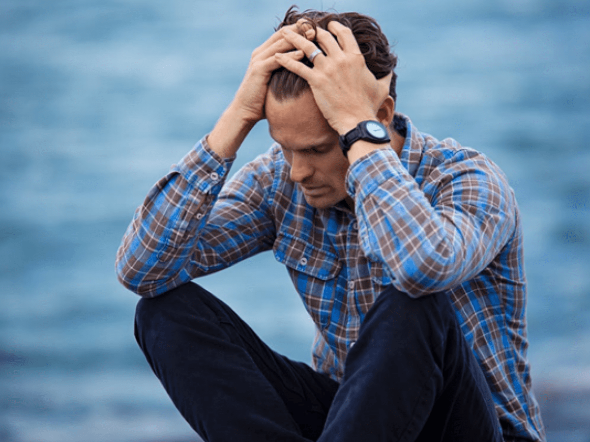 Man wearing blue plaid shirt with both hands on his forehead, looking distressed.