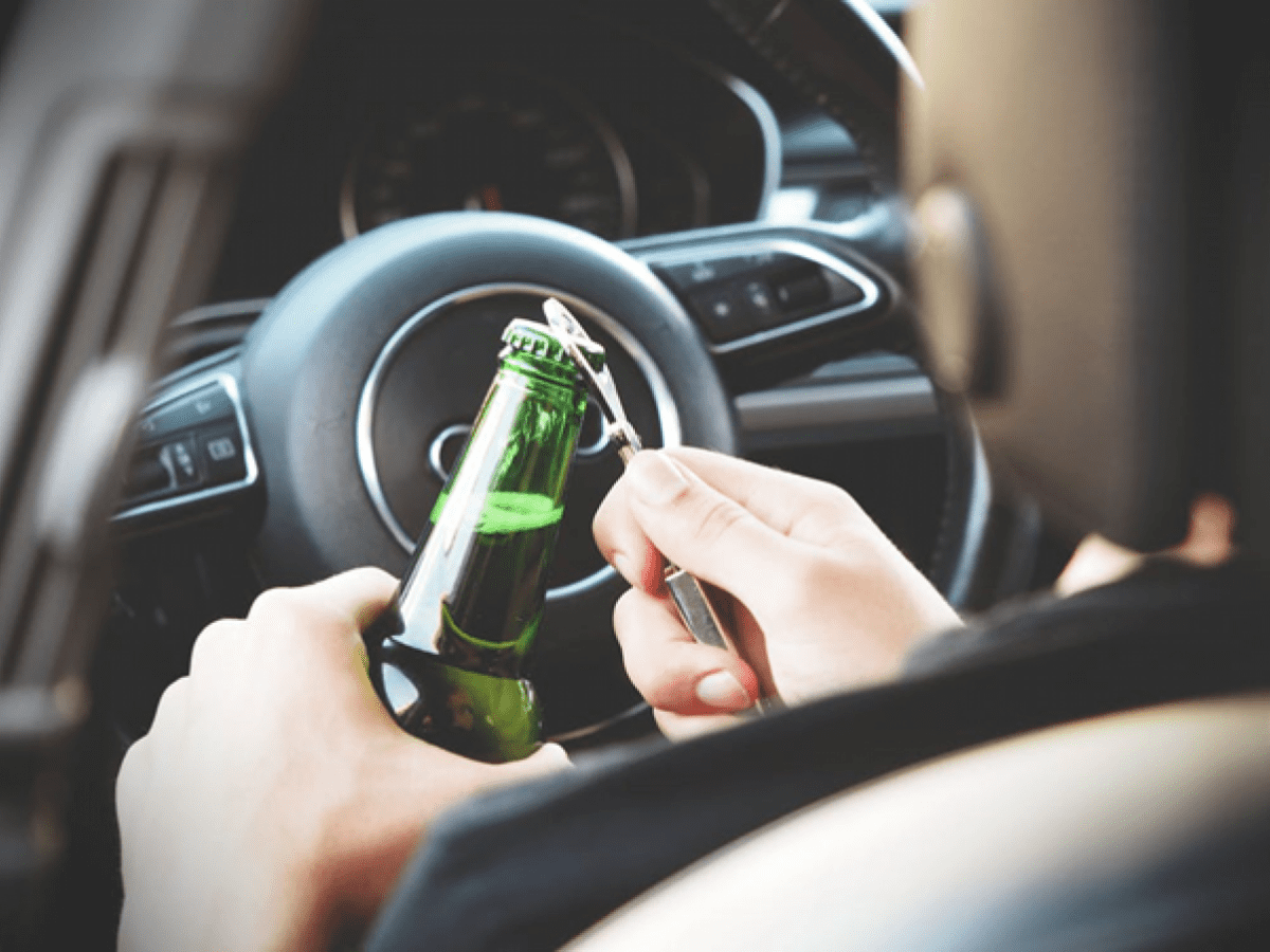 A person opening a bottle of beer with a can opener in the drivers seat of a car.